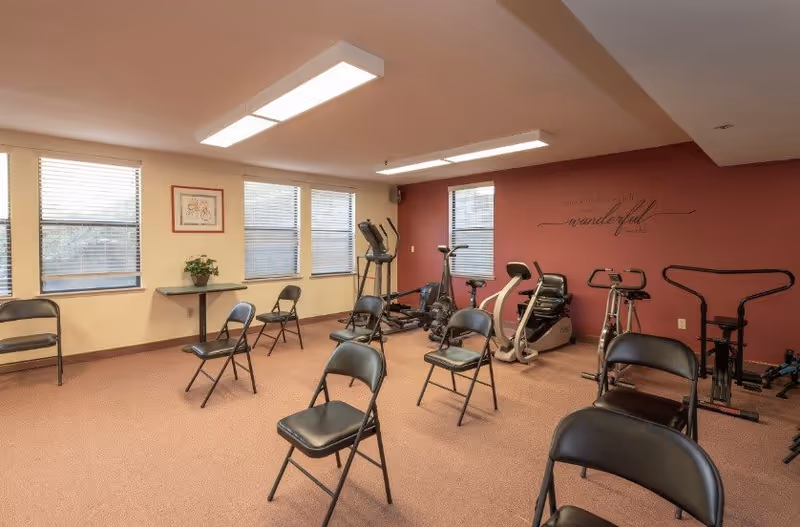 A senior living exercise room with folding chairs arranged in rows and several cardio machines against a red accent wall.