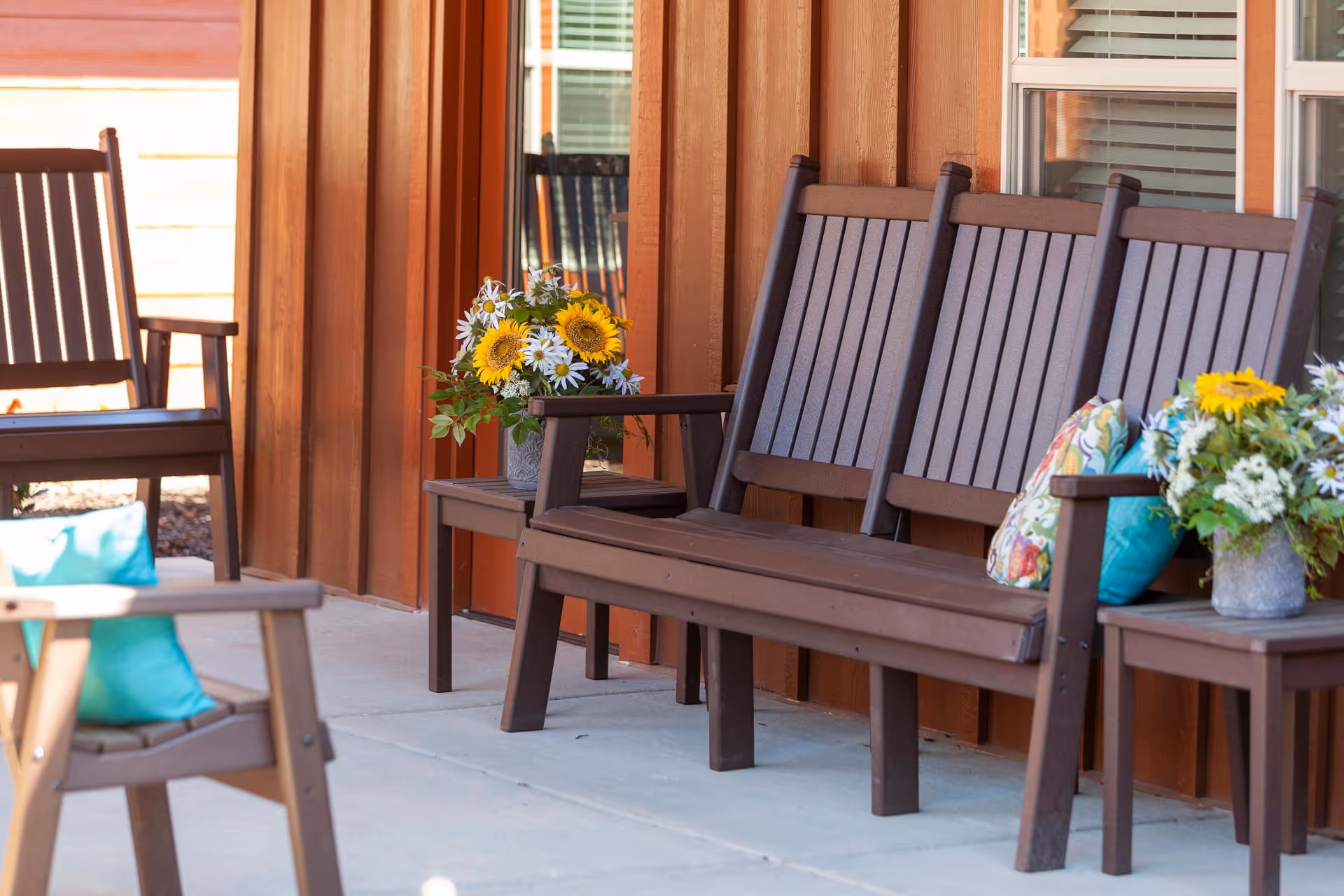Wooden outdoor porch with brown benches and chairs, colorful pillows and potted flowers against a rust-colored siding.