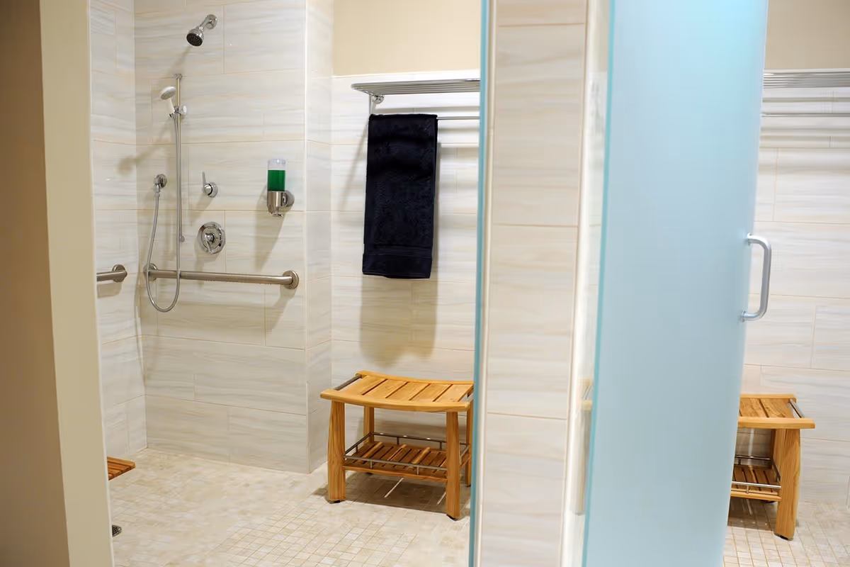 A clean, accessible shower area with beige tiled walls and floor, featuring a wall-mounted showerhead, a handheld showerhead, a horizontal grab bar, a soap dispenser with green soap, a wooden bench, and a towel rack with a dark towel. A frosted glass door with a metal handle is partially visible.