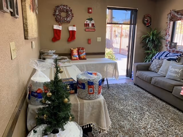 A cozy living room decorated for Christmas with a small decorated Christmas tree on a table, two tables covered with beige tablecloths holding snacks and paper plates, a gray couch with pillows including one that says 'Beyond blessed', Christmas stockings and wreaths hanging on the wall, and a door leading outside letting in natural light.