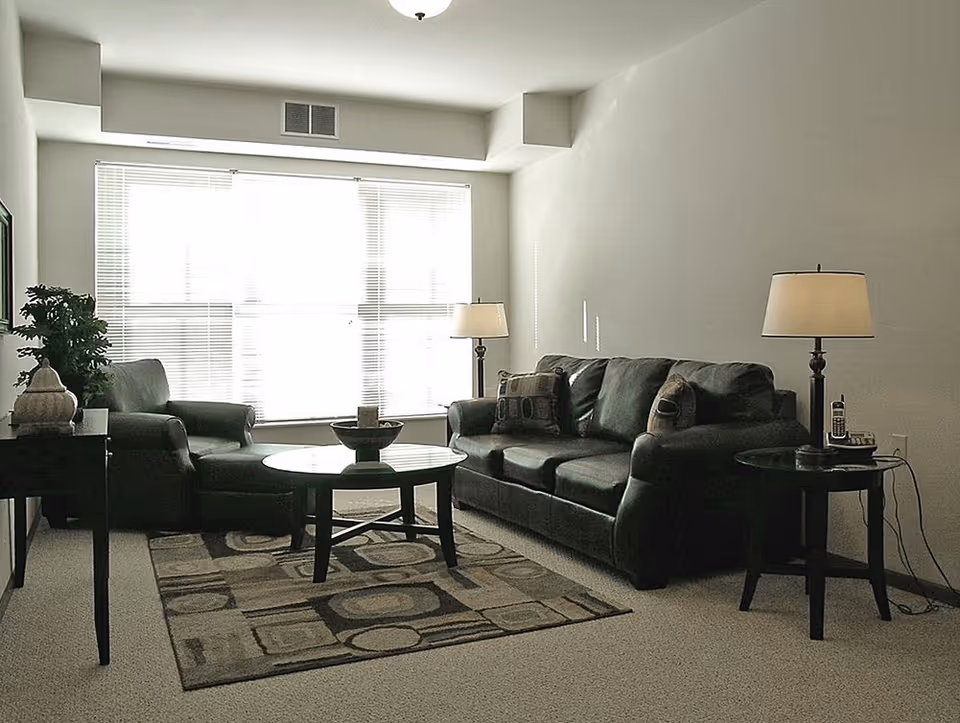 A living room with a large window covered by blinds letting in bright light. The room features a black leather sofa with two patterned pillows, a matching black leather armchair with an ottoman, a round coffee table with a decorative bowl, two side tables each with a lamp, and a patterned area rug on a carpeted floor. There is also a small plant on a side table near the armchair.