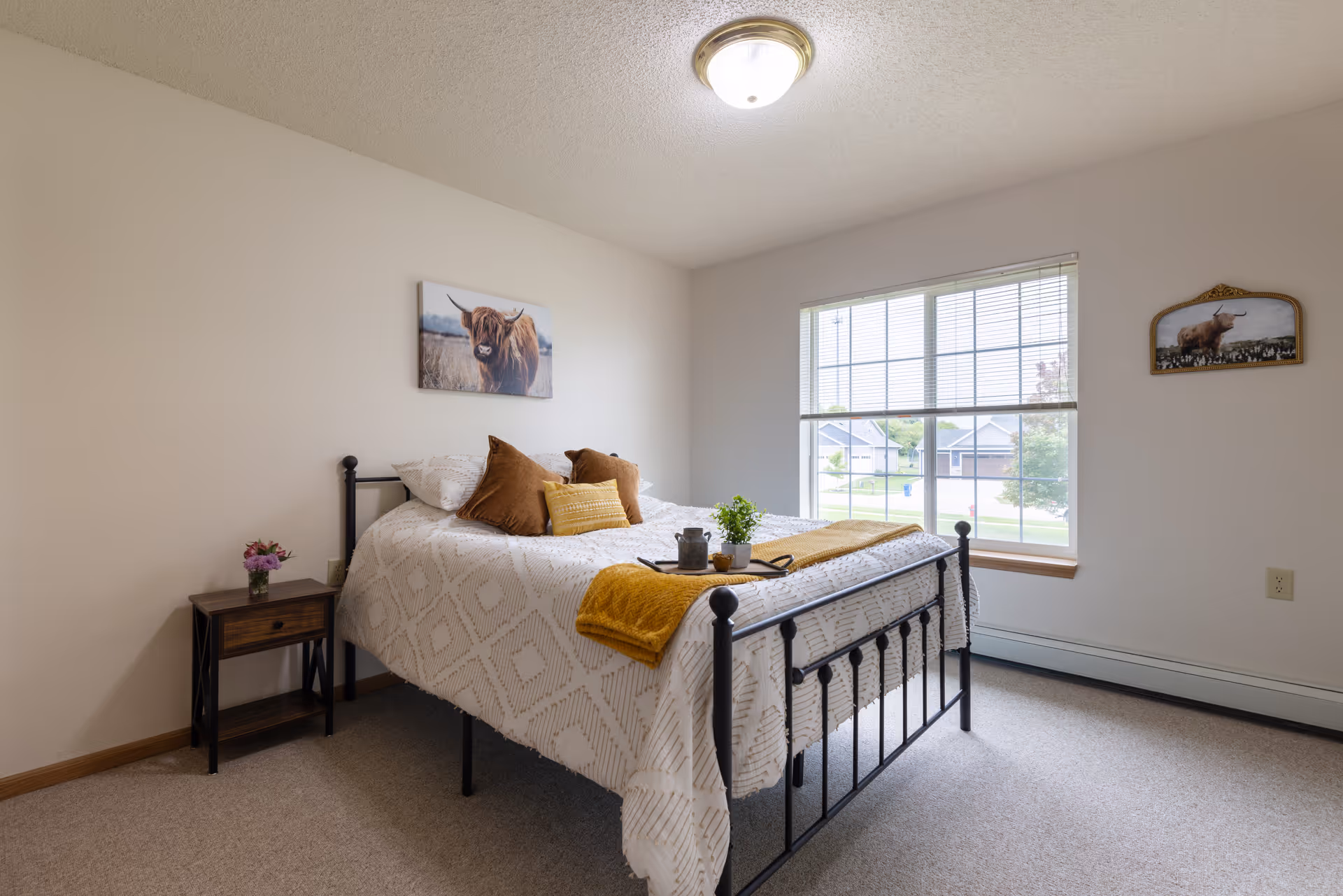 A bright bedroom with a black metal bed frame, white patterned bedding, and brown and yellow accent pillows. A small wooden nightstand with a vase of flowers is beside the bed. Two paintings of a highland cow hang on the walls. A large window with blinds lets in natural light and shows a suburban neighborhood outside.