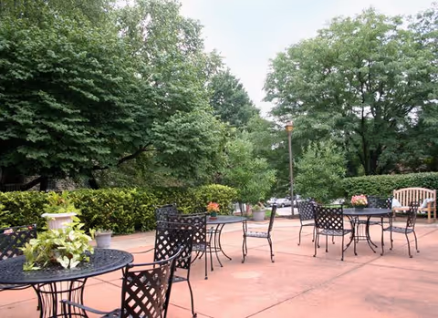 Outdoor patio with metal tables and chairs surrounded by trees and landscaped shrubs.