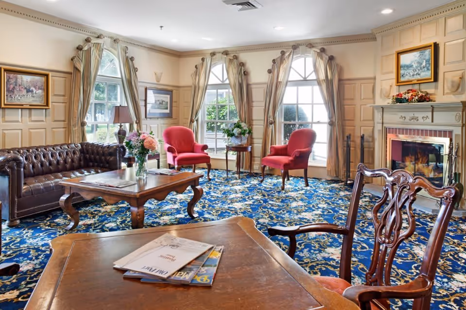 A cozy living room with a blue patterned carpet, a brown leather tufted sofa, two red upholstered armchairs, a wooden coffee table with a vase of flowers, and a fireplace with a floral arrangement on the mantle. Large windows with beige curtains allow natural light to fill the room, and framed paintings hang on the walls.