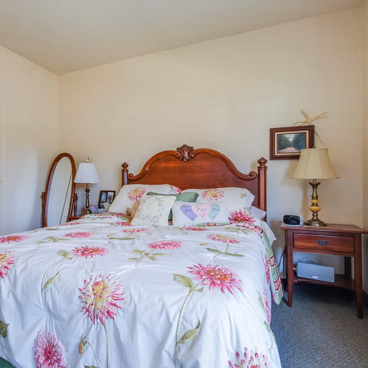 A cozy bedroom featuring a wooden headboard, floral bedspread, bedside tables with lamps, and a floor mirror.