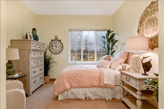 A cozy bedroom with a large window letting in natural light. The bed is made with pink and white bedding and several pillows. On the right side of the bed is a nightstand with a decorative lamp and a small sculpture. On the left side of the room is a tall dresser with decorative items on top and a round wall clock above a potted plant. The walls are painted a soft beige color.