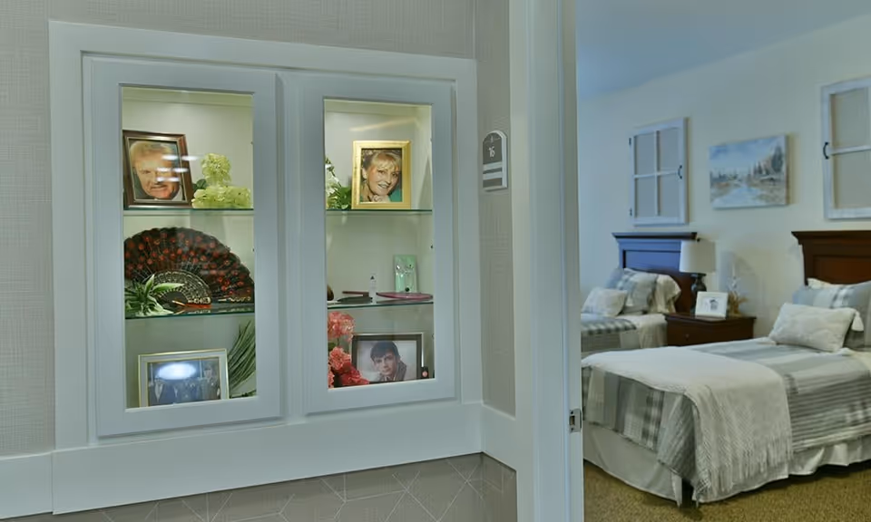 A cozy bedroom with two twin beds, each with gray and white bedding and pillows. Between the beds is a wooden nightstand with a lamp and a framed photo. The walls are decorated with framed artwork and window-like wall decor. In the foreground, there is a built-in glass display cabinet with framed photos, plants, and decorative items.