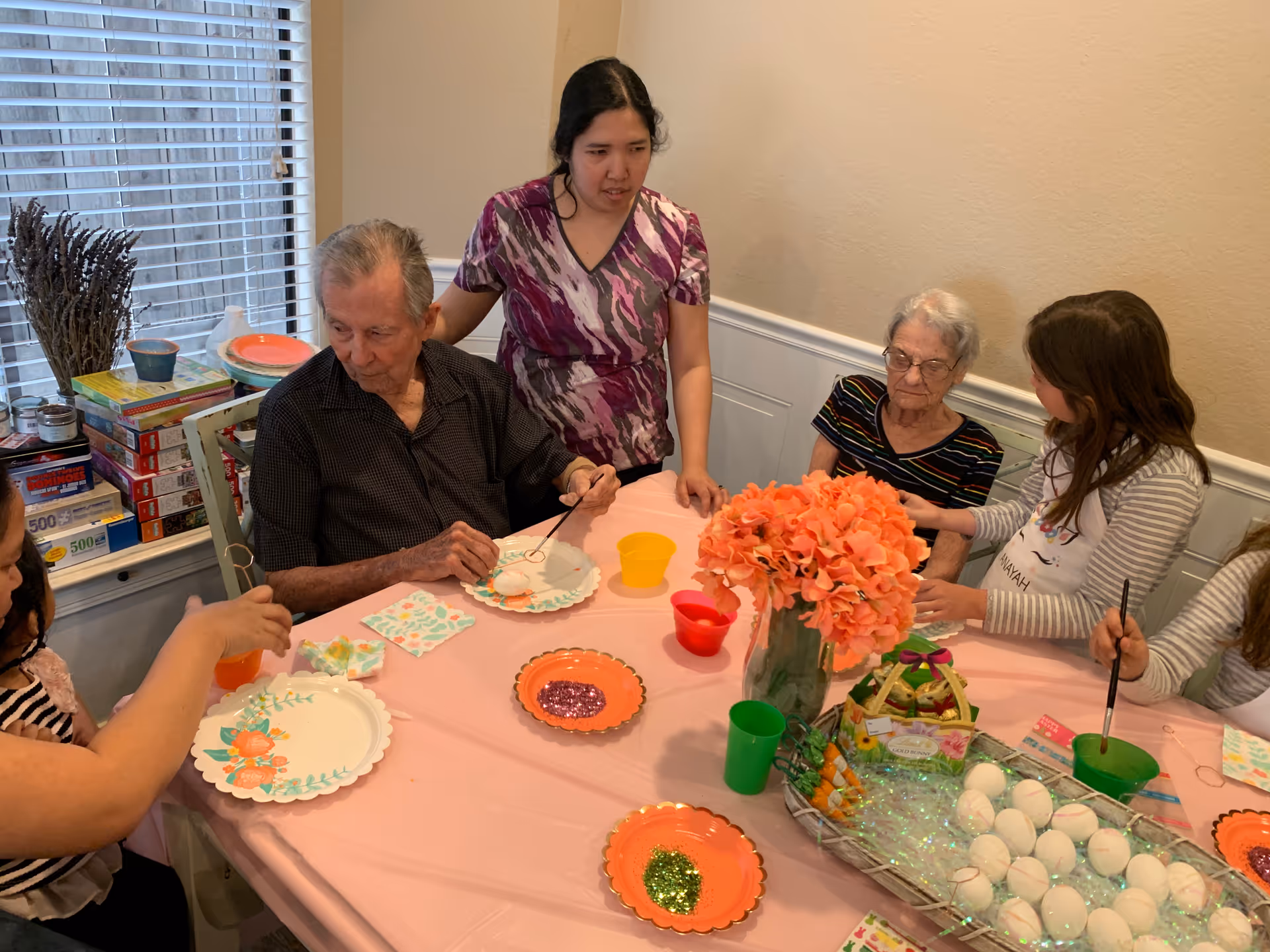 A group of elderly and younger people sitting around a table covered with a pink tablecloth, engaged in an arts and crafts activity involving painting eggs. There are plates with glitter, cups, and a basket of white eggs on the table, along with a vase of orange flowers. The setting appears to be a cozy indoor room with a window and blinds in the background.