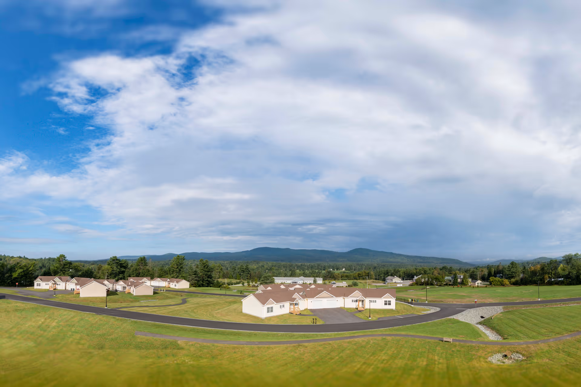 Aerial view of a senior living facility named Summit by Morrison, showing multiple single-story buildings with beige walls and brown roofs, surrounded by green lawns and paved roads, with a backdrop of forested hills and a partly cloudy blue sky.