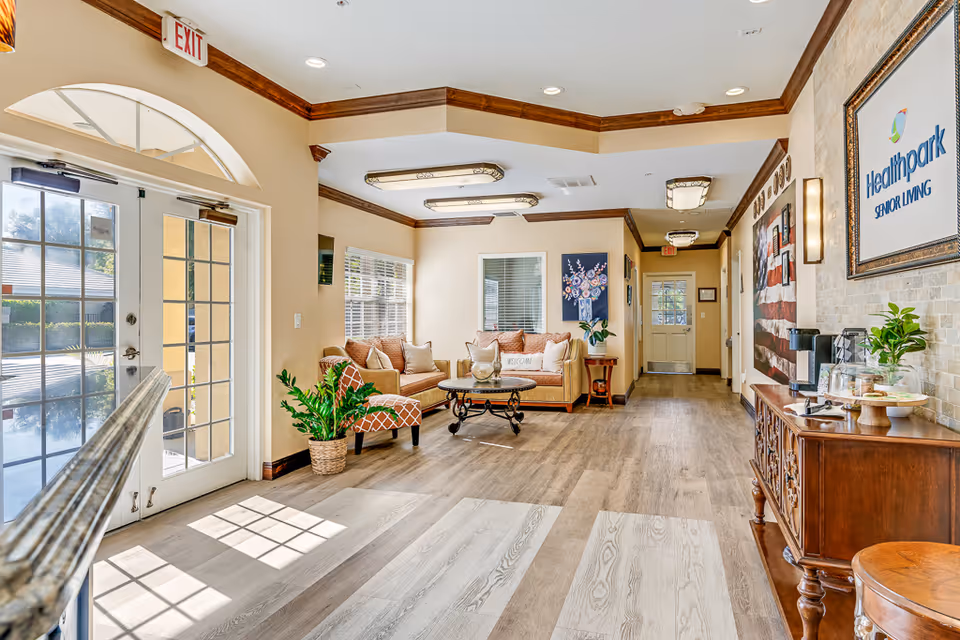 Bright and welcoming senior living facility lobby with wooden flooring, beige walls, and crown molding. The space features a seating area with a sofa, armchair, and coffee table, a potted plant, and a side table with a flower vase. On the right side, there is a wooden console table with a coffee station and snacks. A framed sign on the wall reads 'Healthpark Senior Living'. Large windows and glass doors let in natural light, and exit signs are visible above the doors.