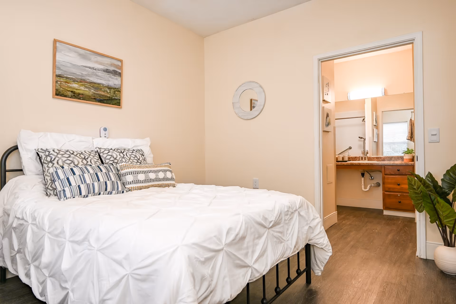 A cozy bedroom with a neatly made bed featuring white bedding and multiple decorative pillows. Above the bed hangs a landscape painting. A round mirror is mounted on the beige wall to the right. The room has wooden flooring and a doorway leading to a bathroom with a sink, mirror, and wooden cabinets. A potted plant is placed on the floor near the bathroom entrance.