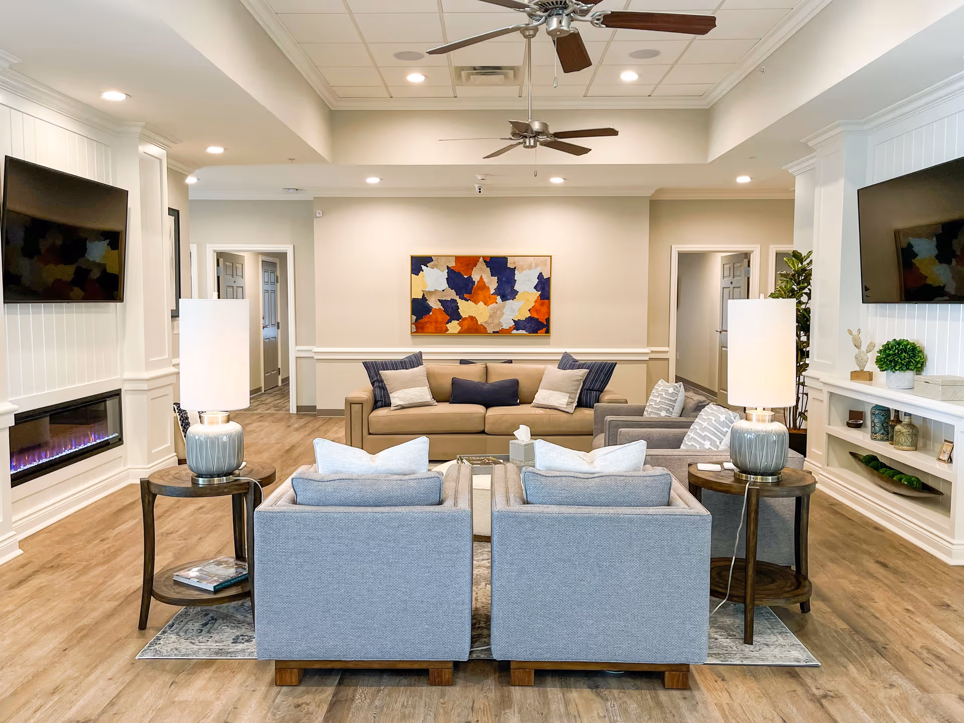 A bright and spacious senior living facility common area with a beige sofa, two gray armchairs, two side tables with lamps, a wall-mounted TV, an electric fireplace, and colorful abstract artwork on the wall. The room has wood flooring, ceiling fans, and recessed lighting.