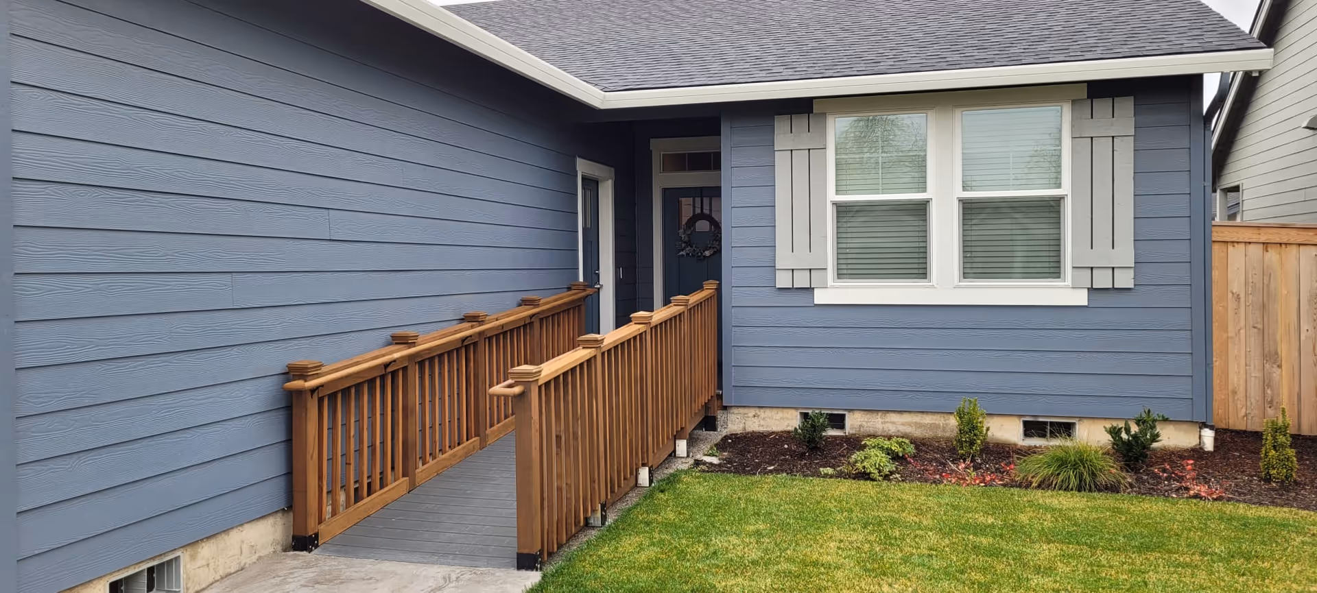 Exterior view of a blue house with a wooden ramp leading to a door. The house has white-trimmed windows with shutters and a small landscaped area with grass and plants.