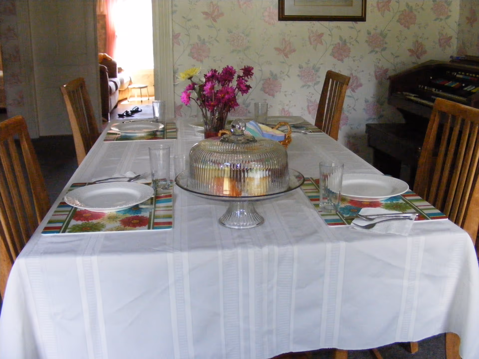 A dining table set for four with white plates, glasses, and silverware on floral placemats. A glass cake stand with a dome cover containing pastries is in the center, along with a vase of purple and yellow flowers. The room has floral wallpaper and a wooden organ is visible in the background.