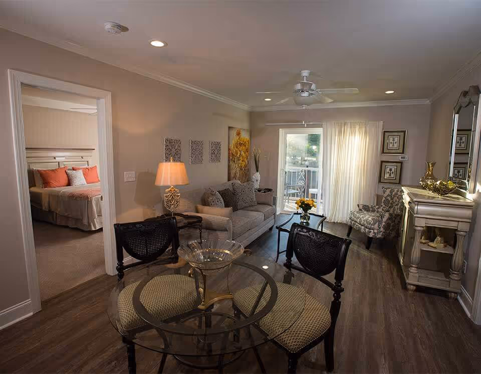 A cozy living room with a gray sofa, patterned armchair, glass-top dining table with four chairs, a lamp on a side table, and a decorative console table with a mirror. The room has wood flooring, recessed ceiling lights, and a ceiling fan. A sliding glass door with sheer curtains leads to an outdoor balcony. To the left, an open doorway reveals a bedroom with a bed featuring coral and white pillows.