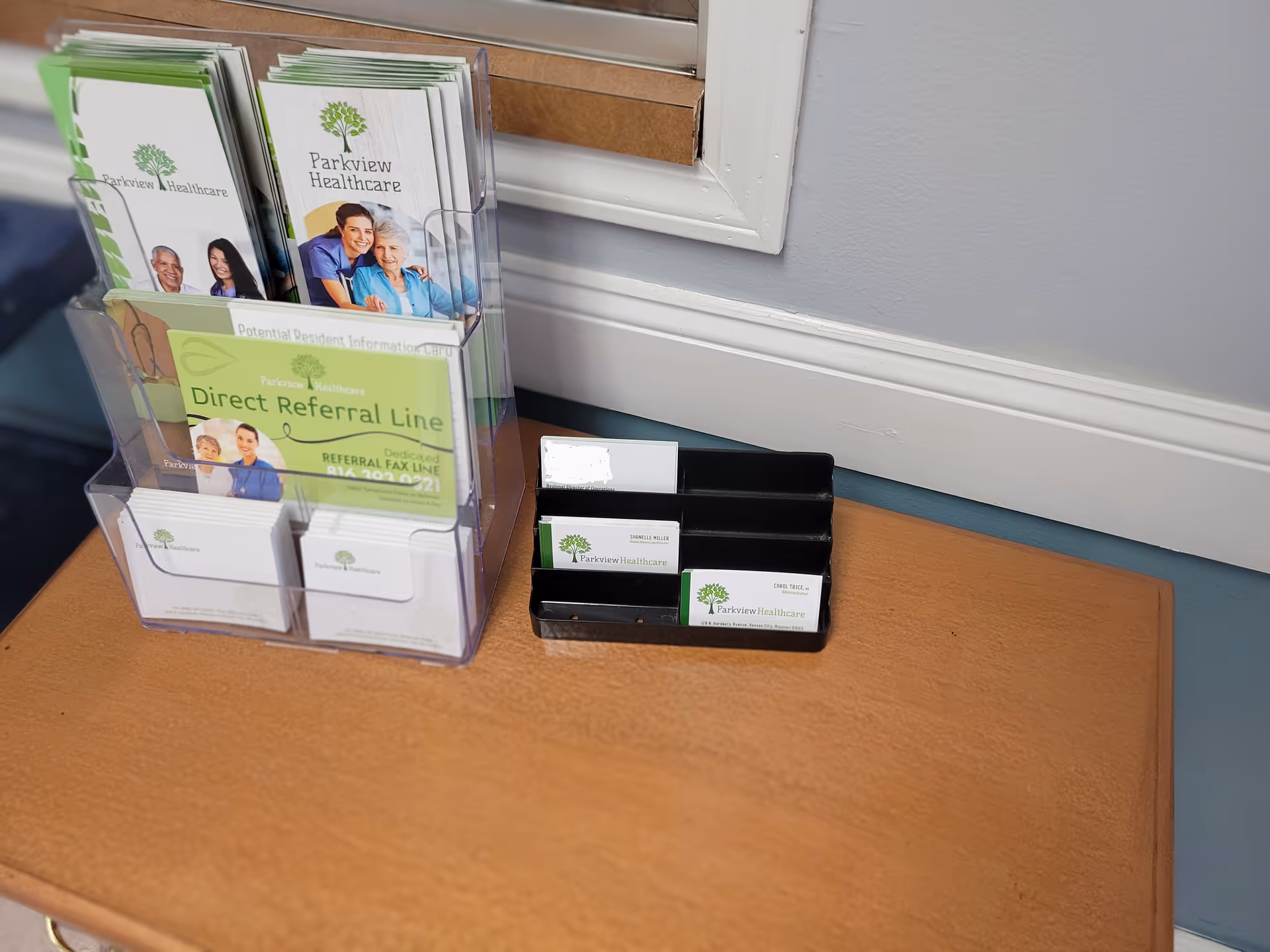 A wooden table with a clear plastic brochure holder containing Parkview Healthcare brochures and a black business card holder with Parkview Healthcare business cards, placed against a gray wall with white trim.