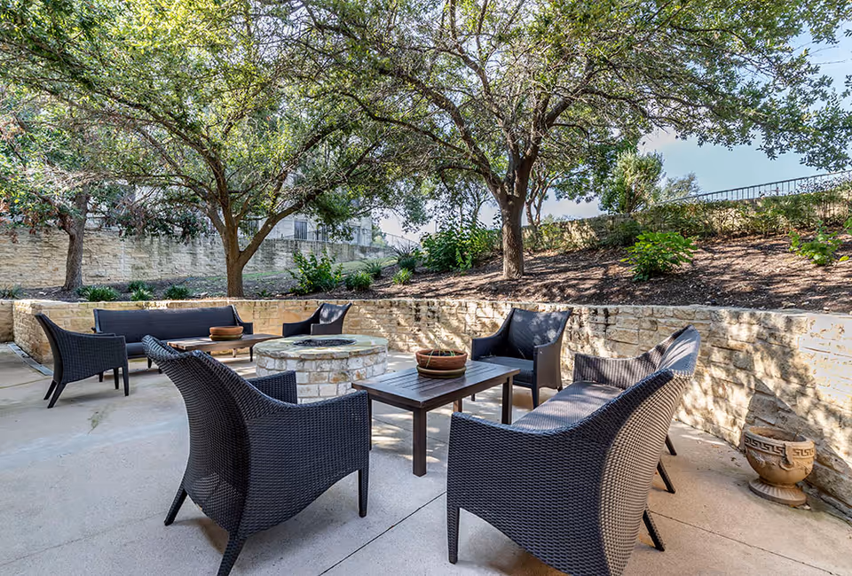 Outdoor patio with wicker seating arranged around a stone fire pit beneath trees and a stone retaining wall.