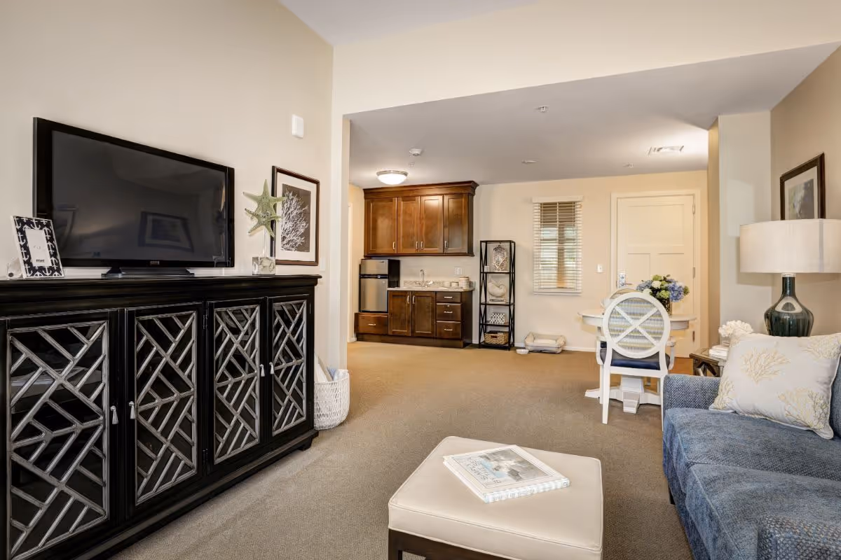 Cozy living room with a TV on a decorative cabinet, a blue sofa and ottoman, and a small kitchenette and dining table in the background.