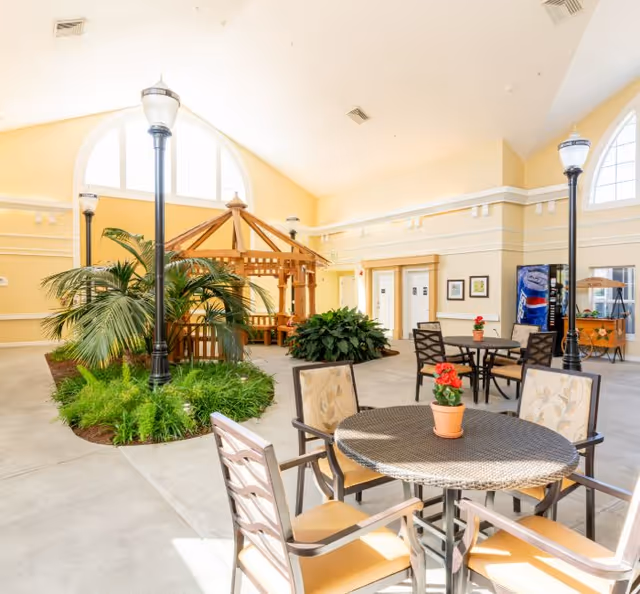 Bright indoor atrium with round tables and chairs, potted plants, a wooden gazebo, and tall streetlamp-style lights.