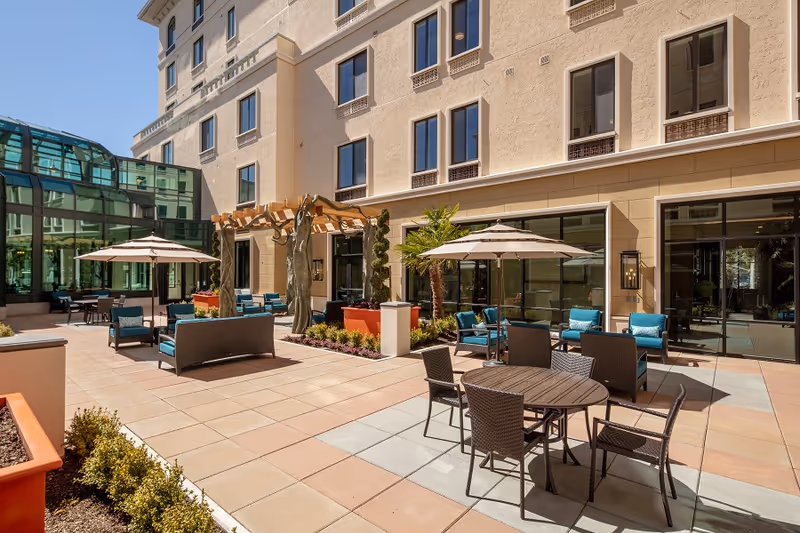 Outdoor patio area at a senior living facility with multiple seating arrangements including round tables with umbrellas and cushioned chairs. The patio is adjacent to a multi-story building with large windows and a glass-enclosed sunroom or atrium on the left side. There are decorative plants and a wooden pergola structure in the center.