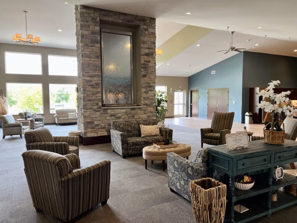 A spacious and well-lit assisted living facility common area with multiple upholstered chairs and sofas arranged around a central stone fireplace. Large windows allow natural light to fill the room. A teal console table with decorative items and a basket of white flowers is positioned in the foreground.