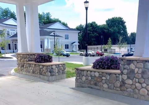 Covered entrance with stone walls and columns, purple flowering planters beside a residential building exterior.