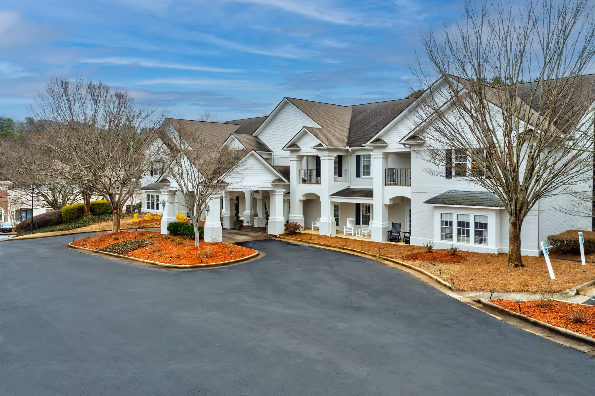 Exterior view of a two-story white building with a covered entrance and balconies, surrounded by leafless trees and landscaped areas with mulch, under a partly cloudy sky.