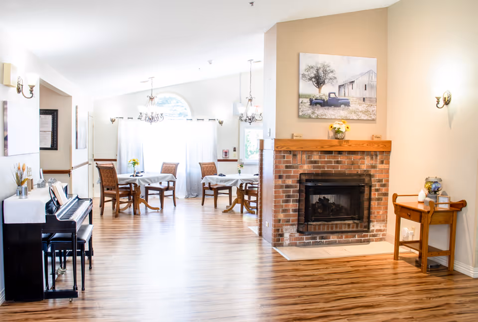Bright communal dining and lounge area with hardwood floors, a brick fireplace, piano, and several dining tables and chairs.