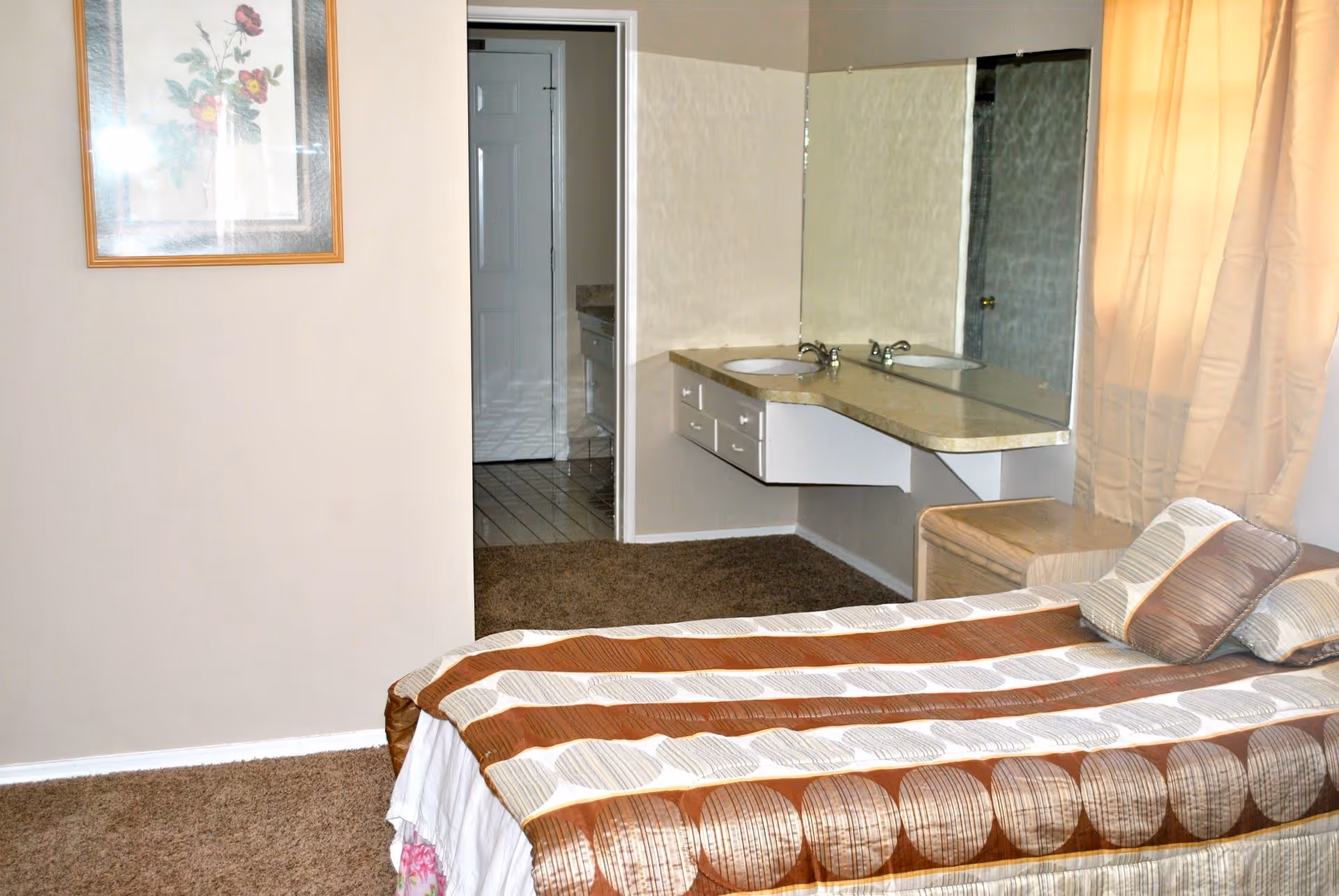 A bedroom in a senior living facility featuring a single bed with a patterned brown and beige bedspread and matching pillows. There is a vanity area with a countertop, two sinks, and a large mirror. A framed floral picture hangs on the wall, and a window with beige curtains allows natural light into the room. The floor is carpeted, and a doorway leads to an adjacent bathroom area with tiled flooring.