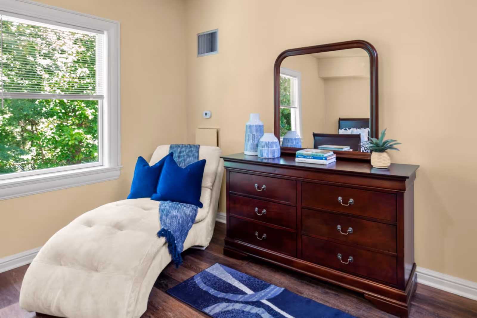 A cozy corner of a room featuring a beige chaise lounge with two blue pillows and a blue throw blanket. Next to it is a dark wooden dresser with six drawers and a large mirror on top. The dresser holds decorative vases, books, and a small potted plant. A window with white blinds shows green foliage outside. The floor is wooden with a blue patterned rug in front of the dresser.