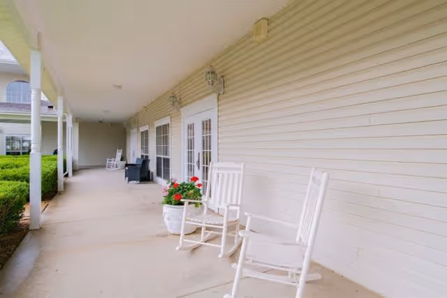 Covered outdoor porch area with white rocking chairs, potted flowers, and a view of the building exterior with windows and doors.