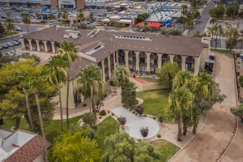 Aerial view of Olive Grove senior living facility showing a large two-story building with a brown roof surrounded by palm trees and landscaped green areas. There are paved walkways and a parking lot visible in the background.