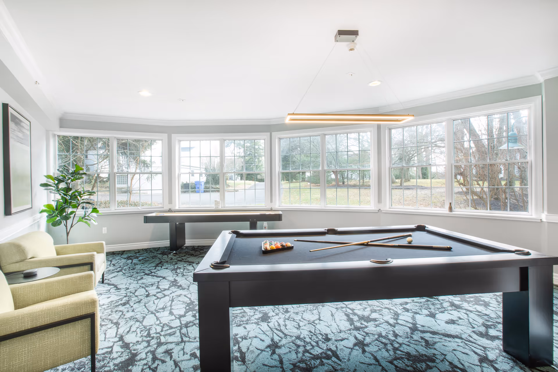 Bright recreational room with large windows showing outdoor greenery, featuring a black pool table with pool cues and balls arranged, a shuffleboard table in the background, two light green armchairs, a potted plant, and a modern hanging light fixture.