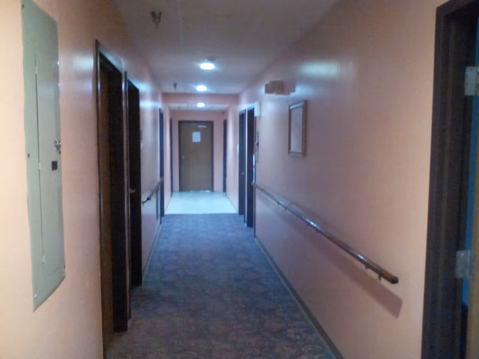 A long indoor hallway with peach-colored walls, carpeted floor, handrails on both sides, several doorways along the corridor, and ceiling lights illuminating the space.