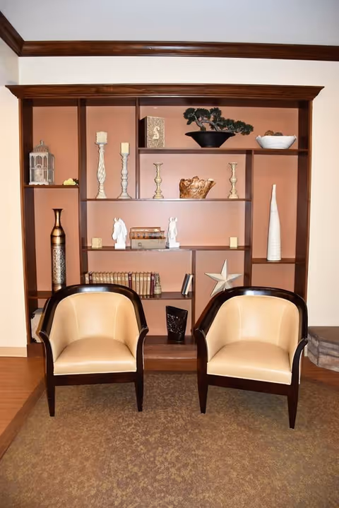 Two beige and dark wood armchairs placed in front of a wooden built-in bookshelf filled with decorative items such as candlesticks, books, a star ornament, a bonsai tree, and vases in a warmly lit room.