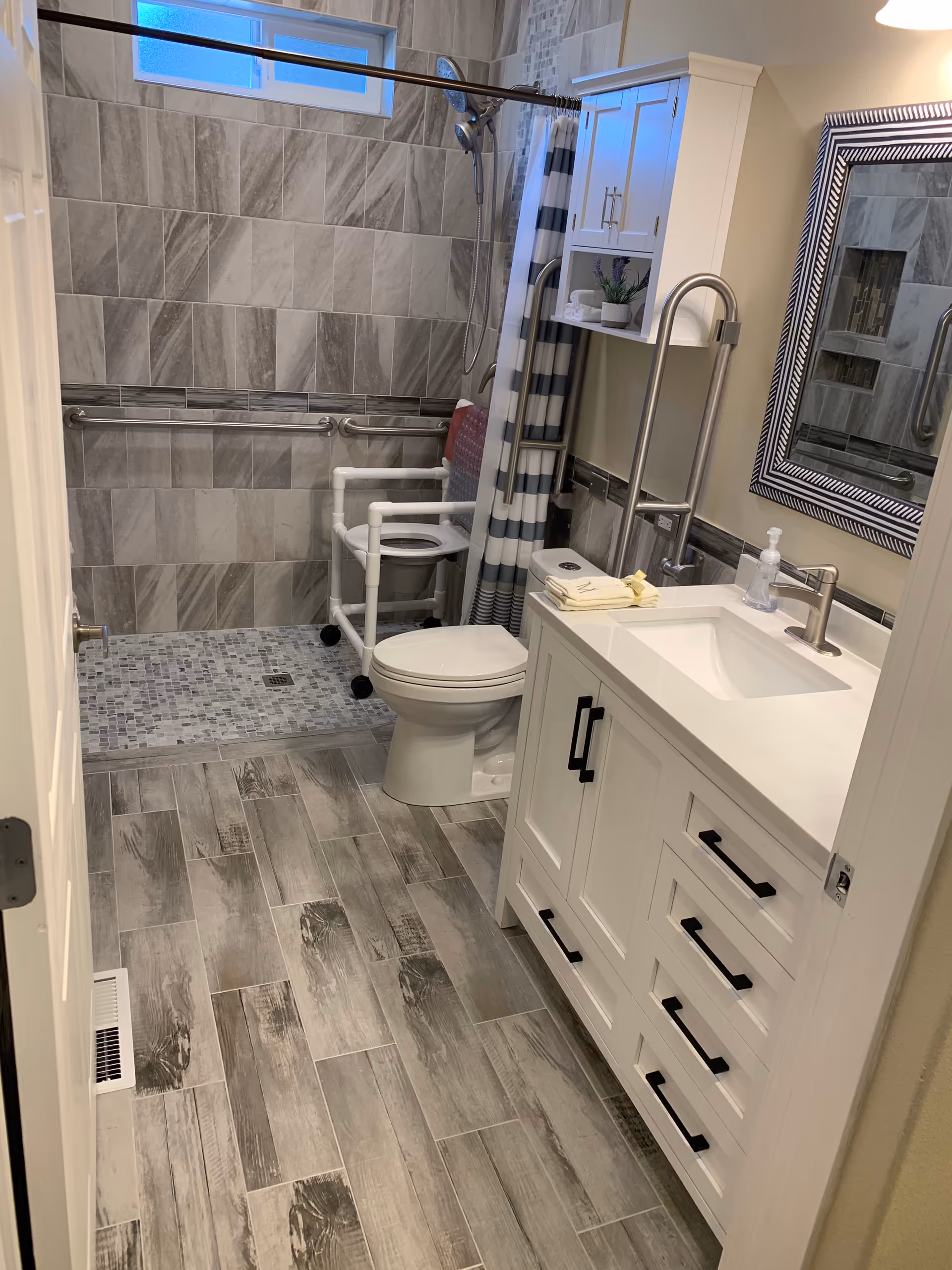 A modern bathroom with gray tiled walls and floor. There is a walk-in shower area with a handheld showerhead and a striped shower curtain. A white toilet is next to the shower, equipped with a white safety frame with armrests. A white vanity with black handles holds a sink, a soap dispenser, and folded towels. A large mirror with a decorative frame hangs above the sink. The bathroom has grab bars for accessibility and a small window near the ceiling.