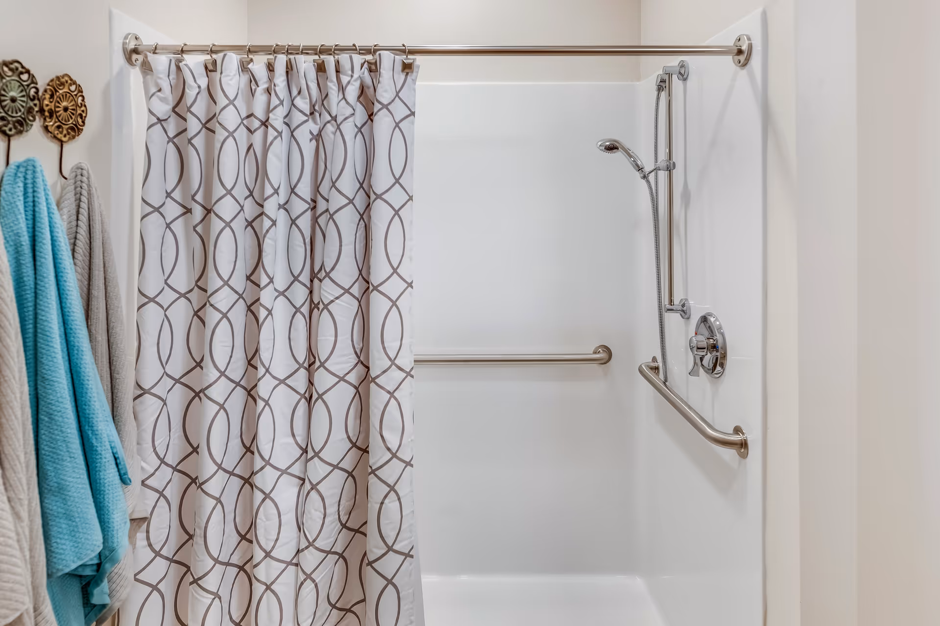 A bathroom shower area with a white and brown patterned shower curtain partially drawn. The shower has a handheld showerhead and two stainless steel grab bars installed on the walls. On the left side, there are three towels hanging on decorative hooks, one turquoise and two beige.