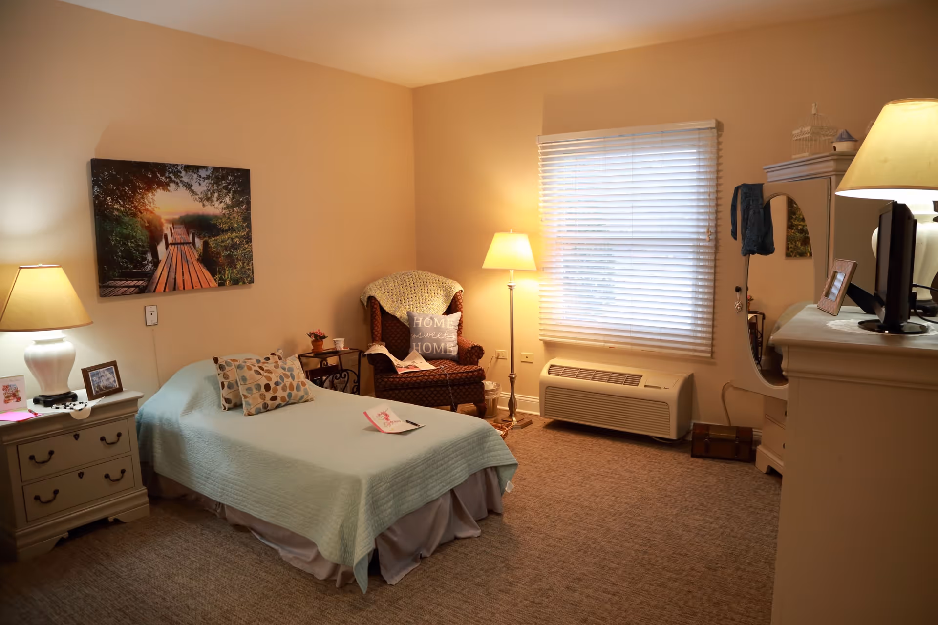 Cozy senior bedroom with a twin bed, armchair, lamps, dresser and a window with blinds.