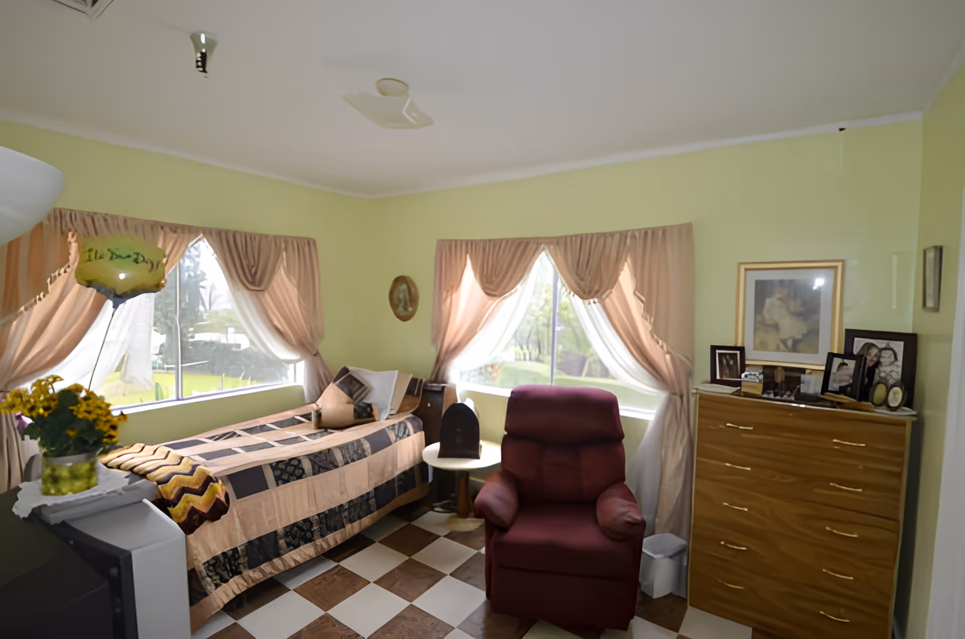 A cozy bedroom with light green walls and two large windows with pink curtains letting in natural light. The room features a single bed with a patterned quilt, a burgundy recliner chair, a wooden dresser with framed photos and artwork on top, and a small round side table. There is a vase with yellow flowers and a balloon on a piece of furniture near the bed.