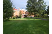 A large green lawn with trees on both sides leading up to a multi-story brick building under a partly cloudy sky.