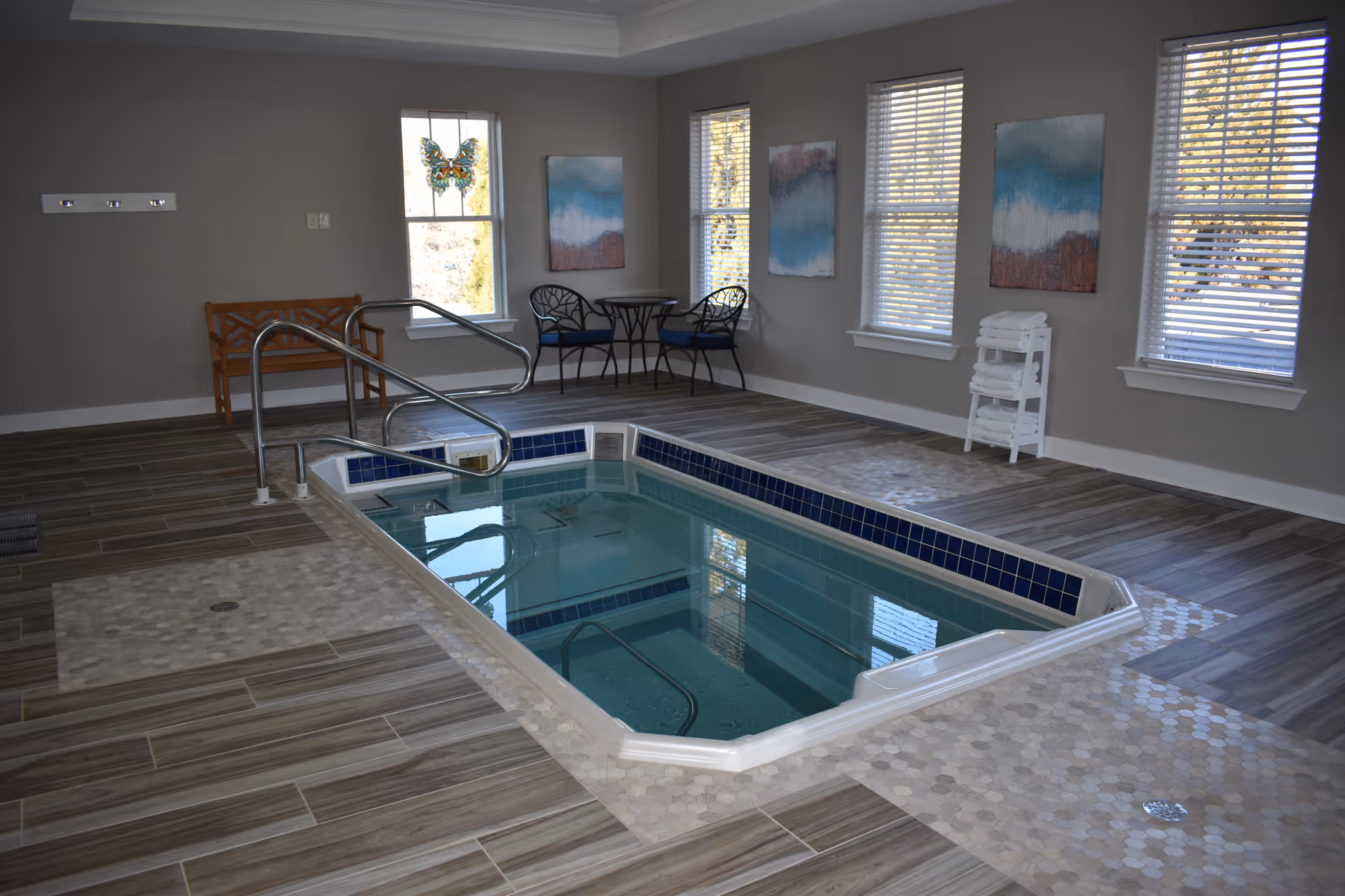 Indoor therapy pool in a bright room with windows, tiled floor, metal handrails, chairs and a towel rack.