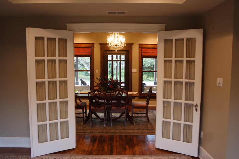 Open white French doors reveal a formal dining room with a wooden table and chairs beneath a chandelier and windows overlooking a garden.