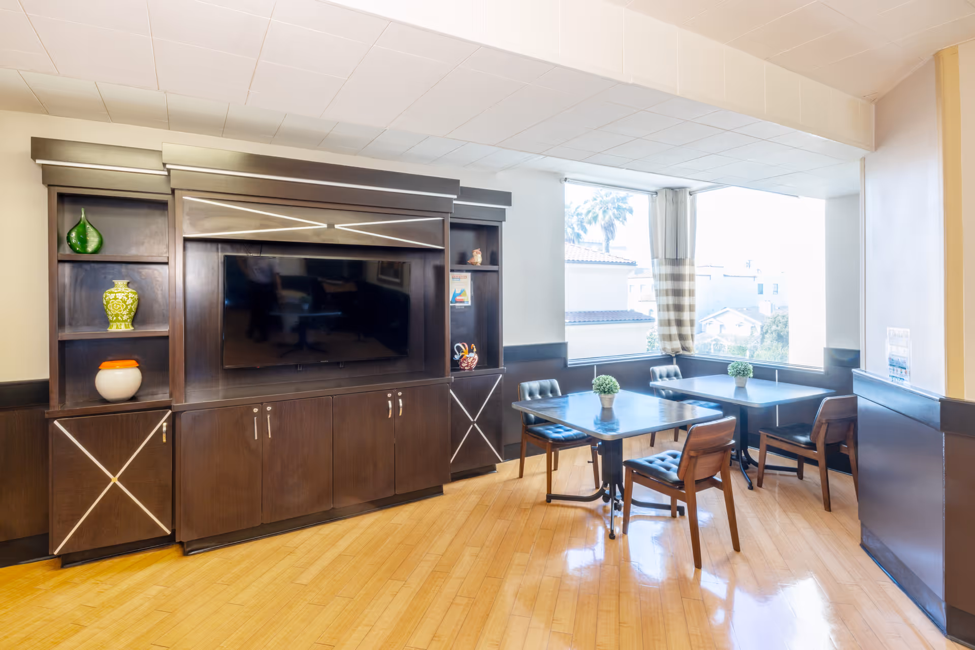 A bright room with large windows featuring two tables each surrounded by four chairs. There is a dark wooden entertainment center with a flat-screen TV mounted in the middle and decorative vases on the shelves. The floor is light wood, and small potted plants are placed on the tables.