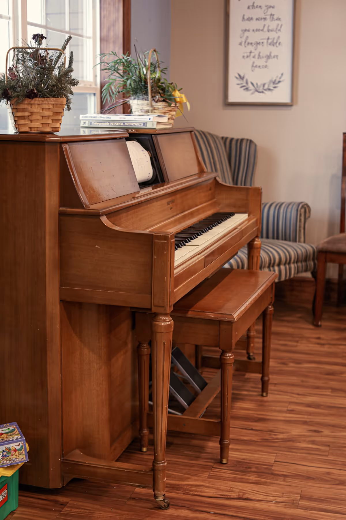 A wooden upright piano with a matching bench in a room with wooden flooring. On top of the piano are two baskets with plants and a few books. In the background, there is a striped armchair and a framed wall art with cursive writing.