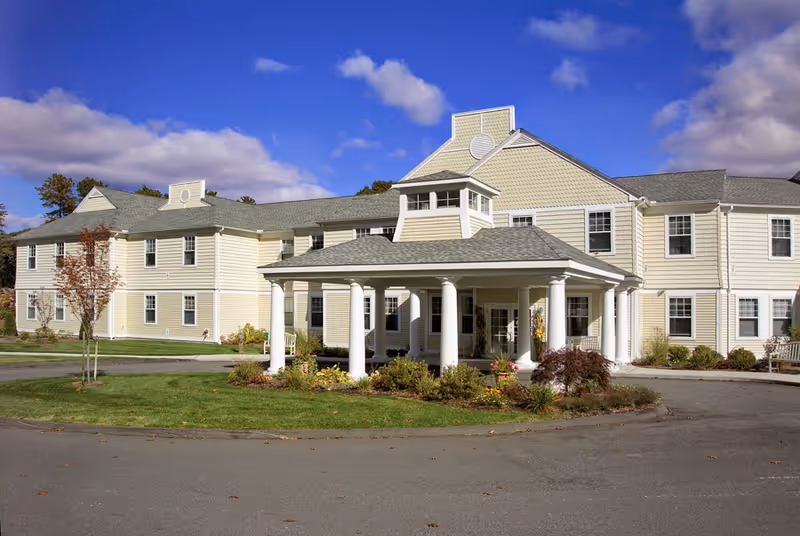 Front exterior of a two-story beige senior living building with a columned entrance portico and circular driveway under a blue sky.