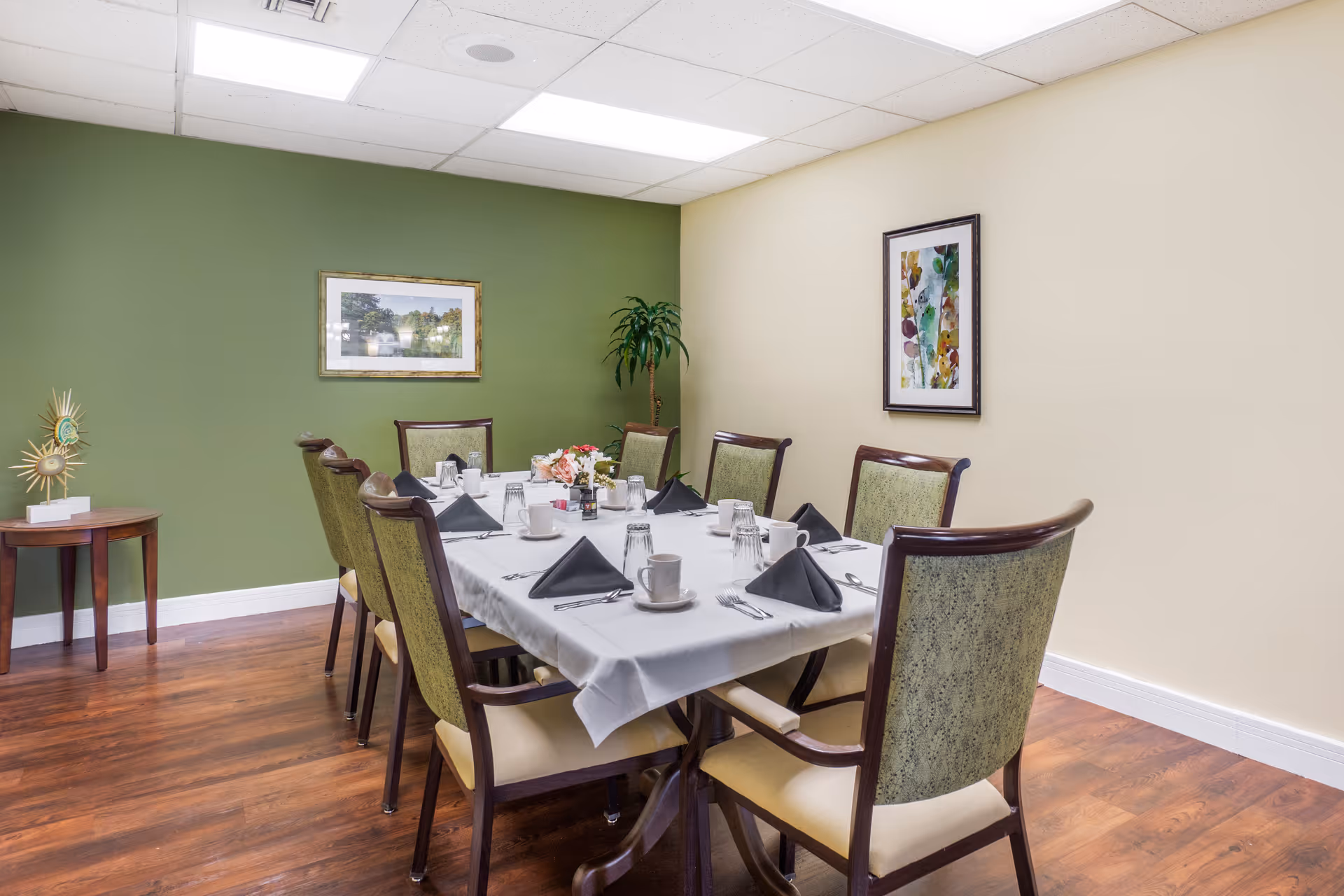 A dining room with a rectangular table set for eight people. The table is covered with a white tablecloth and has black folded napkins, cups, glasses, and silverware arranged neatly. The room has wooden flooring, green and beige walls, two framed pictures on the walls, and a small side table with decorative items.