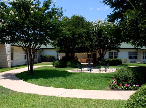 Well-maintained courtyard with a circular paved walkway, green lawn, trees, a small fountain and outdoor seating in front of a single-story building.