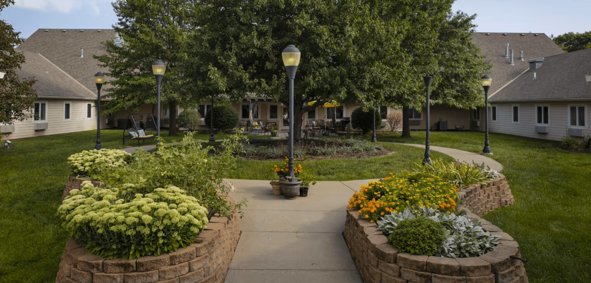 A landscaped outdoor courtyard area at Homestead Assisted Living of Shawnee featuring a paved walkway flanked by raised flower beds with green and yellow plants. Several lamp posts line the walkway, and there are trees and shrubs surrounding the area. The courtyard is enclosed by single-story buildings with beige siding and multiple windows.