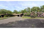 Exterior view of a single-story nursing and rehabilitation center building with a covered entrance driveway. The building is surrounded by greenery, including trees and flowering bushes, under a partly cloudy sky.