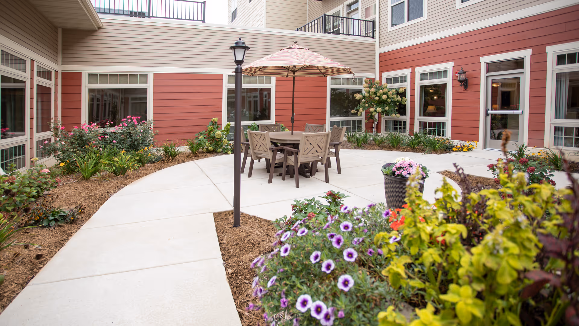 Outdoor courtyard area at a senior living facility with a concrete pathway, garden beds with various flowers and plants, a table with chairs and an umbrella, and red and beige building walls with multiple windows and a door.
